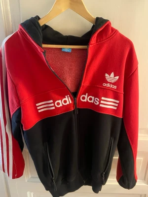 Röd och svart Adidas hoodie med dragkedja - Snygg hoodie från Adidas i rött och svart med vit logga och tryck framtill. Klassiska tre ränder längs ärmarna och dragkedja hela vägen. Utrustad med huva och två fickor med dragkedja. Mjuk fleece på insidan för extra komfort.