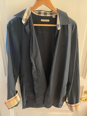 Gammal Svart skjorta från Burberry - Stilren svart skjorta från Burberry med klassiskt rutigt mönster på insidan av krage och manschetter. Skjortan har långa ärmar, knappar framtill och är tillverkad i mjuk bomull. Perfekt för dig som gillar en tidlös look med en twist.