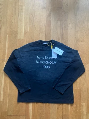 Svart långärmad topp Acne Studios longsleeve - Cool svart långärmad topp från Acne Studios med vit text 'Acne Studios STOCKHOLM 1996' framtill. Klassisk rund halsringning och relaxed fit. Perfekt för dig som gillar minimalistisk och stilren design.