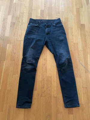 Slim fit jeans från nudie jeans - Snygga svarta slim fit jeans med diskreta slitningar på knäna och lätt tvättad look. Jeansen har klassisk femficksdesign och smal passform som sitter tajt längs benen. Perfekta för en trendig och avslappnad stil.