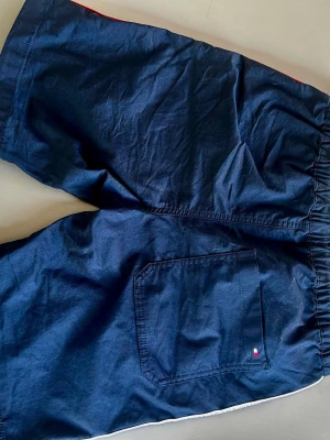 Blå shorts från Tommy Hilfiger - Säljer ett par mörkblå shorts från Tommy Hilfiger med elastisk midja och snygg logotypdetalj längs kanten. Shortsens material är bomull och de har en bakficka med liten Hilfiger-flagga. Perfekta för sommarens chill dagar.