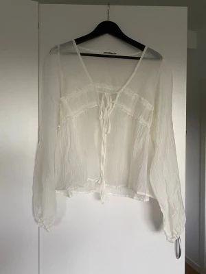Vit transparent blus med knytband - Säljer en luftig och transparent vit blus med långa ärmar och knytband framtill. Blusen har en romantisk känsla med volangdetaljer och är tillverkad i ett tunt, skrynkligt tyg som ger en avslappnad look.