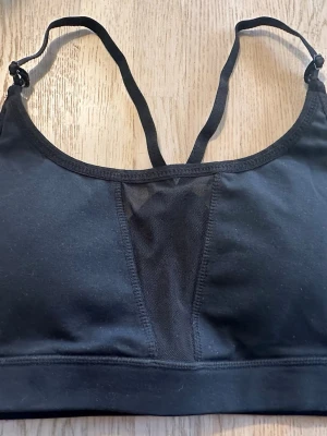 Svart sport-bh med meshdetaljer - Snygg svart sport-bh från Better Bodies med tunna, justerbara axelband som korsas i ryggen. Bh:n har meshdetaljer framtill och baktill för extra ventilation och en bred resår under bysten för bra stöd. Perfekt för träning och aktiva dagar. Ny men utan lapp