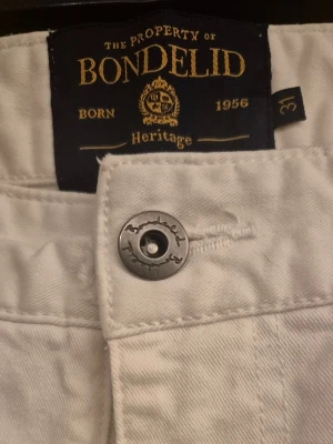 Vita herr jeans från Bondelid Heritage - Snygga vita jeans från Bondelid Heritage med klassisk femficksdesign och raka ben. Jeansen har en stilren knapp med Bondelid-logga och är tillverkade i ett mjukt bomullsmaterial. Perfekta för dig som vill ha en clean och fräsch look. Knappen har gått sönder en gång men är lagad. 