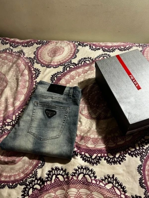 Blåa jeans från Prada - Snygga blåa jeans från Prada med klassisk femficksdesign och diskret tvättad look. Jeansen har raka ben och en svart patch med logga baktill samt Pradas ikoniska trekantslogga på bakfickan. Perfekta för dig som gillar exklusiva märken och stilren design.