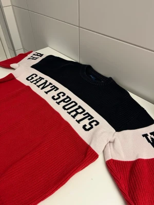 GANT Sportswear stickad tröja - Stickad tröja från GANT med breda färgblock i marinblått, vitt och rött. Tröjan har rund halsringning och texten 'GANT SPORTSWEAR' i svart över bröstet. Perfekt för dig som gillar sportig och klassisk stil.