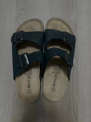 Svarta sandaler med spännen - Klassiska svarta  sandaler med två breda remmar och justerbara metallspännen. Fotbädden är i mocka och har en naturlig beige ton. Perfekta för en avslappnad och trendig look under varma dagar.