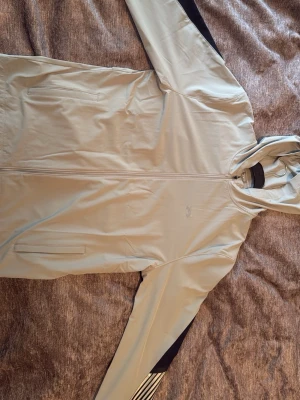 Ljusgrå vindjacka från Under Armour - Ljusgrå vindjacka med huva från Under Armour. Jackan har svarta och vita detaljer på ärmarna, hel dragkedja framtill och två sidofickor med dragkedja. Materialet är lätt och funktionellt, perfekt för att skydda mot vind. Snygg och sportig design.