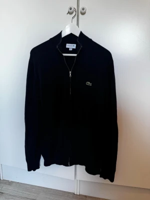 Marinblå ziptröja från Lacoste - Klassisk marinblå ziptröja från Lacoste med ståkrage och dragkedja framtill. Tröjan har det ikoniska krokodilmärket broderat på bröstet och är i en skön classic fit-modell. Perfekt till lager på lager och enkel att matcha med olika outfits.