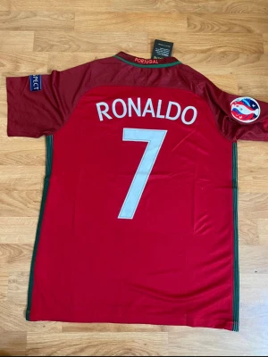 Portugal Ronaldo 7 matchtröja Nike - Snygg röd Portugal fotbollströja 2016 från Nike med Ronaldo och nummer 7 på ryggen. Tröjan har officiella märken, gröna detaljer längs sidorna och patchar på ärmarna. Perfekt för dig som älskar fotboll och vill stötta Portugal.