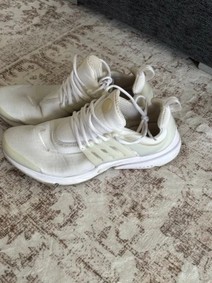 Vita Nike Presto sneakers - Säljer ett par fräscha vita Nike Presto sneakers med mesh-ovandel, vita snören och plastdetaljer på sidorna. Skorna har en låg silhuett och en bekväm, sportig design som passar perfekt till streetwear. Ikonisk Nike-logga på tån och plös.