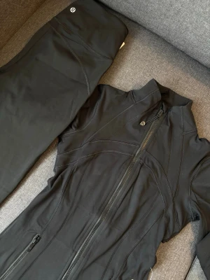 Svart träningsset från Lululemon - Snyggt svart träningsset från Lululemon med figursydd jacka och matchande byxor. Jackan har dragkedja, hög krage och två fickor med dragkedja. Byxorna är utsvängda och har diskret logga. Perfekt för gymmet eller yoga. Storlek är M men passar perfekt till S .Priset kan diskuteras 💖🫶🏻labels are not available in the items 🫶🏻