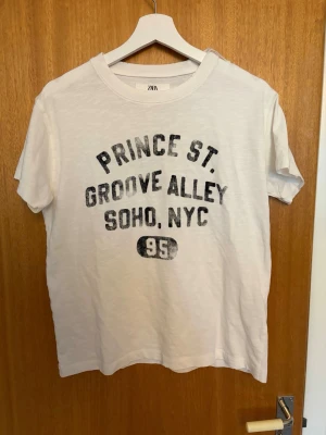 Vit t-shirt med tryck  - Vit t-shirt från ZARA med svart vintageinspirerat tryck på bröstet där det står 'PRINCE ST. GROOVE ALLEY SOHO, NYC 95'. Klassisk rund halsringning och korta ärmar. Tillverkad i mjuk bomull för en skön och avslappnad look. Helt ny med prislapp kvar.