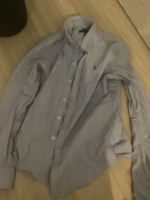 Ljusblå skjorta från Ralph Lauren - Klassisk ljusblå skjorta från Ralph Lauren med broderad logga på bröstet. Skjortan har lång ärm, knappar framtill och en tidlös design som passar till många olika stilar. Tillverkad i bomull för en skön känsla. Passar för både tjejer och killar!!
