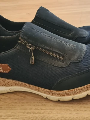 Mörkblå skor med dragkedja - Snygga mörkblå loafers från Rieker med dragkedja på ovansidan och detaljer i textil och mocka. Skorna har en vit och brun sula med sportig känsla och en liten läderdetalj på sidan. Perfekta för en avslappnad stil.