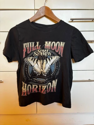 Svart t-shirt med fjärilstryck från Gina Tricot storlek XS - Svart t-shirt med tryck framtill 'Full Moon Horizon Soul Search' och en stor fjäril i mitten. T-shirten har rund hals och korta ärmar. Från Gina Tricot storlek XS