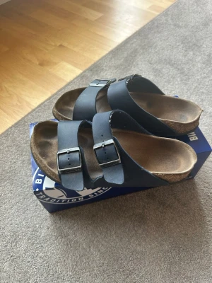 Svarta Birkenstock sandaler - Klassiska svarta sandaler från Birkenstock med två breda remmar och justerbara metallspännen. Fotbädden är i kork och mocka för extra komfort. Perfekta för en avslappnad och trendig stil under varma dagar.
