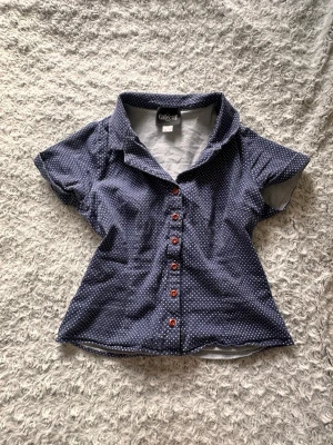 Blå prickig skjorta från Collectif - Kortärmad blå skjorta från Collectif med vita prickar och bruna knappar framtill. Skjortan har klassisk krage och figurnära passform, perfekt för en fest eller till vardags. Tillverkad i mjuk stretchig bomull som känns skön mot huden.