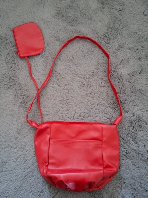 Röd axelväska i konstläder - Cool röd axelväska i mjukt konstläder med enkel design och en matchande liten plånbok. Väskan har en lång axelrem och rynkad botten för en trendig look. Perfekt för dig som vill sticka ut med färg och stil.