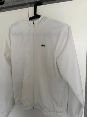 Vit vindjacka från Lacoste - Snygg vit vindjacka från Lacoste med dragkedja framtill och klassisk krokodil-logga på bröstet. Jackan har elastiska muddar vid ärmslut och nederkant samt en minimalistisk design. Perfekt för dig som gillar stilrena plagg med sportig känsla. OBS! 176cm men passar även 168 till cirka 175cm lång