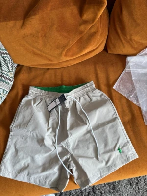 Beige badbyxor från Polo Ralph Lauren - Snygga beige badbyxor från Polo Ralph Lauren med grön insida och klassisk grön logga broderad på benet. De har elastisk midja med vit dragsko, sidofickor och en bakficka. Perfekta för stranden eller poolen.