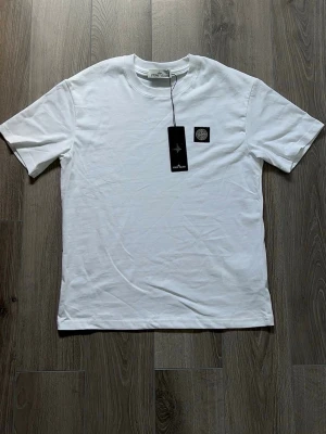 Vit t-shirt från Stone Island - Säljer en clean vit t-shirt från Stone Island med klassisk logopatch på bröstet. T-shirten har korta ärmar och rund halsringning. Tillverkad i mjuk bomull som känns skön mot huden. Perfekt för dig som gillar stilrena plagg med ikonisk detalj.