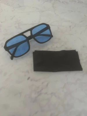 Svarta solglasögon med blåa glas - Snygga solglasögon med svart båge och blåa, fyrkantiga glas. Medföljer en svart putsduk. Perfekta för dig som vill sticka ut med en cool och modern look. Helt nya och oanvända!