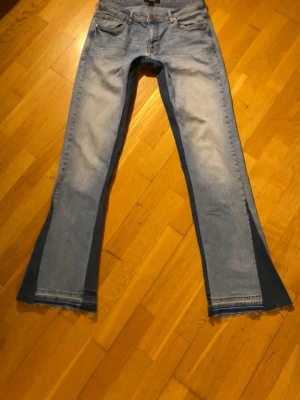 Ljusblå utsvängda jeans med mörka paneler - Säljer ett par ljusblå jeans med utsvängda ben och mörkblå paneler längs sidorna. Jeansen har råa kanter nertill och klassisk femficksdesign. Perfekta för dig som gillar en unik och trendig look. Materialet är denim.