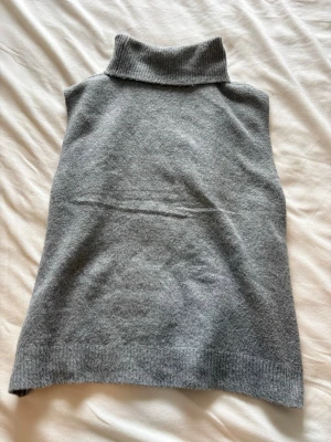 Grå stickad ärmlös polotröja - Säljer en stilren grå stickad ärmlös polotröja med hög ribbad krage. Perfekt att bära över en skjorta eller långärmad topp för en trendig lager-på-lager-look. Mjuk och skön kvalitet, troligtvis i ull eller kashmirblandning.
