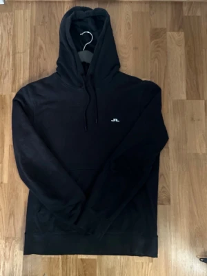 Svart hoodie från J.Lindeberg - Säljer en svart hoodie från J.Lindeberg med liten vit logga på bröstet. Hoodien har huva med dragsko och en klassisk känguruficka framtill. Tillverkad i mjukt material som känns skönt mot huden. Perfekt för en avslappnad och stilren look.