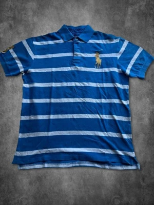 Blåvit randig pikétröja från Polo Ralph Lauren Stl XL - Säljer en blå och vit randig pikétröja från Polo Ralph Lauren med klassisk krage och knappar framtill. Tröjan har ett broderat logomärke på bröstet och en stor guldfärgad siffra 3 på ena ärmen. Tillverkad i mjuk bomull och har korta ärmar.  Mått: Axelbredd: 49cm Pit-to-Pit: 56cm Midja: 54cm Längd: 68cm (räknar inte med kragen)