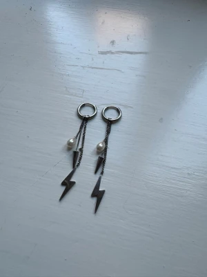 Silvriga blixtörhängen med pärlor - Säljer ett par coola örhängen i silverfärgad metall med hängande blixtar och vita pärldetaljer. Örhängena har ringfäste och tunna kedjor som ger en edgy vibe. Perfekta för dig som vill sticka ut med din stil.