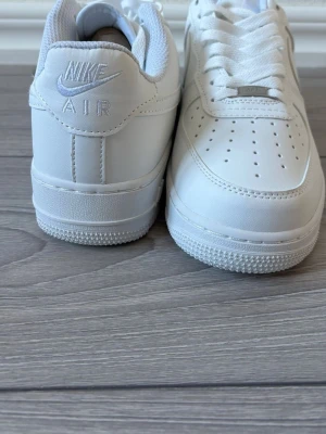 Nike Air Force 1 vita sneakers - Klassiska Nike Air Force 1 sneakers i helvitt läder med perforerad tå, vit sula och ikoniska swoosh-loggan på sidan. Skorna har snörning och AIR-detalj på hälen. Perfekta för en clean och stilren look.