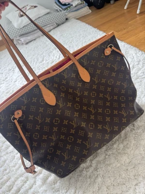 Louis Vuitton Monogram shoppingväska - Säljer en klassisk shoppingväska från Louis Vuitton med ikoniskt monogrammönster i brunt och beige. Väskan har långa läderhandtag och detaljer i ljusbrunt läder. Perfekt för dig som vill ha en rymlig och stilren väska med lyxig känsla.