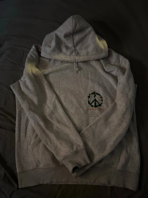 Grå hoodie från H&M - Grå hoodie med huva och känguruficka. Tryck med olika svarta symboler på ryggen och ett mindre tryck på bröstet. Mjuk bomullskänsla och ribbade muddar vid ärmslut och nederkant. Perfekt för en avslappnad stil.