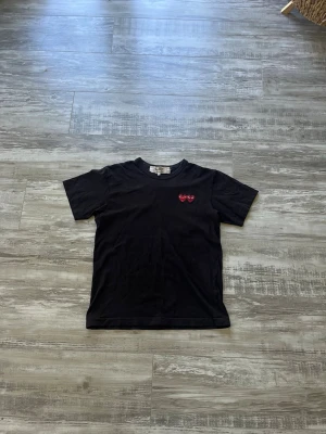 Svart t-shirt med rött hjärta - En svart t-shirt i bomull med korta ärmar och rund hals. På bröstet finns en liten röd hjärtdetalj med ögon, vilket ger en lekfull touch till den annars enkla designen. Perfekt för dig som gillar stilrena plagg med en twist. Tvättas innan den skickas. 