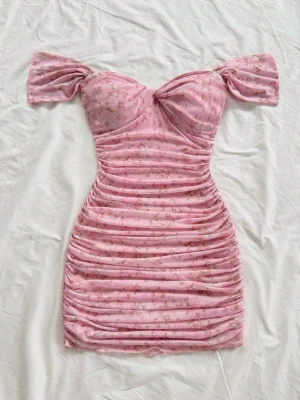 Rosa blommig offshoulder klänning - Söt rosa kortklänning med blommigt mönster och offshoulder-ärmar. Klänningen har rynkade detaljer längs hela kroppen och en feminin, figurnära passform. Perfekt för dig som gillar romantisk och trendig stil.