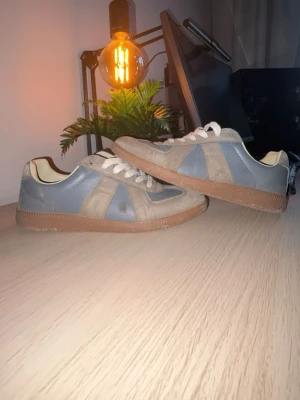 Maison Margiela Gats - Snygga Gats i grått och beige med vita skosnören och bruna gummisulor. Skorna har en klassisk låg profil och kombinerar släta partier i läder med detaljer i mocka för en stilren look. Perfekta för dig som gillar en clean och tidlös stil. Skorna har en defekt (se första bilden) samt är dem lite solblekta. Säljer p.ga dem inte passar mig längre. PRISET ÄR EJ HUGGET I STEN