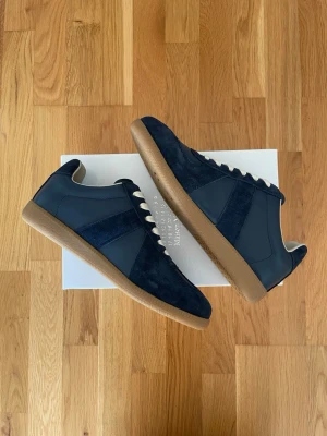 Mörkblå sneakers från Maison Margiela - Snygga mörkblå sneakers från Maison Margiela med detaljer i mocka och släta partier i läderlook. Skorna har vita snören och en klassisk gummisula i beige. Perfekt för dig som gillar stilrena och tidlösa sneakers med en modern twist.