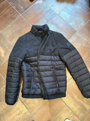 Calvin Klein jacka - Snygg svart pufferjacka med quiltade partier och hög krage. Jackan har dragkedja framtill, två sidofickor med dragkedja och ribbade muddar vid ärmslut och nederkant. Perfekt för kalla vinterdagar och ger en modern look.