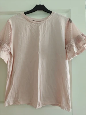 Ljusrosa topp med spetsärmar - Söt ljusrosa topp med korta ärmar i mesh och spetsdetaljer. Ärmarna har prickigt mönster och dekorativ spetskant som ger en romantisk vibe. Perfekt till jeans eller kjol för en trendig look.