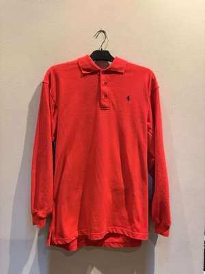 Röd långärmad pikétröja Polo Ralph Lauren - Klassisk röd långärmad pikétröja från Polo Ralph Lauren med krage, knappar vid halsen och den ikoniska broderade loggan på bröstet. Tillverkad i bomull för en bekväm och avslappnad stil.