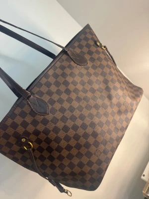 Brun shoppingväska med rutmönster - Snygg shoppingväska i brunt med klassiskt rutmönster och mörkbruna handtag. Väskan har guldiga metalldetaljer och är rymlig med en stilren design. Perfekt för dig som vill ha en trendig och praktisk väska till vardagen. Handtaget är slitet men går fortfarande att använda väskan (se bild nr 3 på defekt)