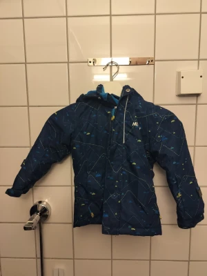Blå vindjacka med mönster från Nevica - Snygg blå vindjacka från Nevica med huva och dragkedja. Jackan har ett diskret mönster i vitt, blått och gult samt justerbara ärmslut. Perfekt för blåsiga dagar och har en sportig look som passar till det mesta.               Storlek: 98-104 cm (3-4 år)
