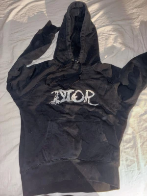 Svart hoodie med tryck - Svart hoodie med stor huva och magficka. På bröstet finns ett vitt tryck i en cool, sliten stil. Hoodien har dragsko vid huvan och är gjord i mjuk bomull. Perfekt för dig som gillar streetwear och vill sticka ut. Ända felet med den är att lappen under luvan är lös på ena sidan men går lätt att sy tillbaka 
