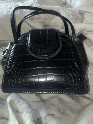 Svart minihandväska i krokodilimitation - Stilren svart handväska med krokodilpräglat mönster och dubbla handtag. Väskan har en rundad lockstängning och detaljer i silver. Perfekt för dig som gillar trendiga accessoarer med lyxig känsla.