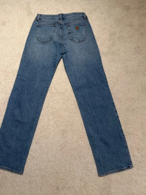 Blå raka jeans med klassisk look - Ett par blå jeans med rak passform och klassisk femficksdesign. Jeansen har en enkel stil med normalhög midja och en liten läderpatch på bakfickan. Perfekta för dig som gillar en tidlös och avslappnad vibe.