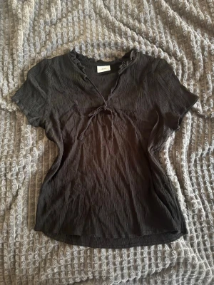 Svart blus med knyt och volang - Säljer en svart blus med korta ärmar, v-ringning och volangkant vid halsen. 