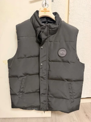 Svart dunväst från canada goose  - Säljer en stilren och varm Canada Goose-väst i svart. Perfekt att använda över en hoodie eller tröja under vår, höst och mildare vinterdagar.  Skick:  * bra  skick * Väl omhändertagen * Inga hål eller större skador * Alla knappar och dragkedjan fungerar som de ska  Detaljer:  * Färg: Svart * Två framfickor * Canada Goose-logga på bröstet * Varm och bekväm passform * Passar både till vardags och finare tillfällen