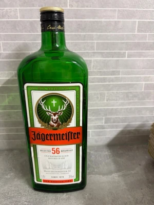 Jägermeister 1,75L grön glasflaska - Stor grön glasflaska Jägermeister med klassisk etikett och orange logga. Flaskan rymmer 1,75 liter . Etiketten visar hjorthuvud och text om 56 örter och kryddor.